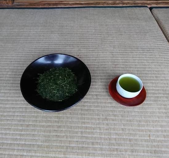 畳の上、黒い漆の皿に盛られた茶葉と、朱の茶托に置かれた一杯の緑茶